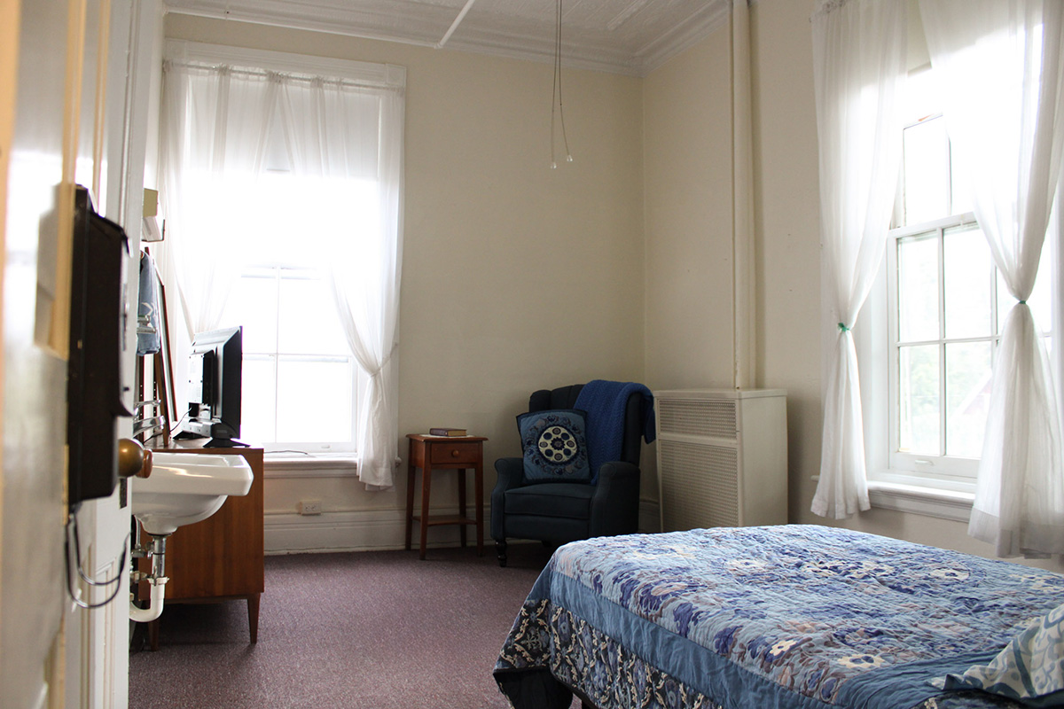 A bedroom at Vassar-Warner home, with a sitting area, sink, television set, and sunlight filtering in through large windows.