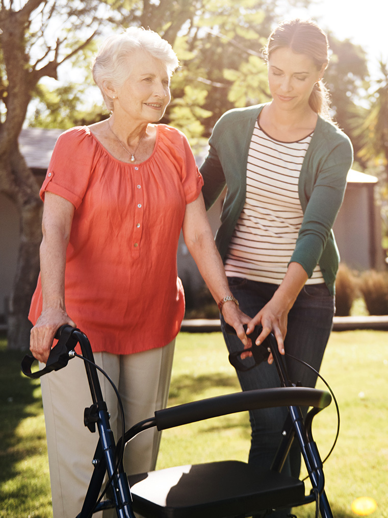 A volunteer assists a senior living resident with her walker while on a stroll through a park on a sunny day.